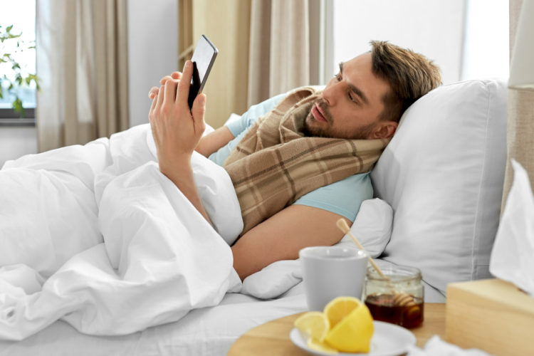 health, illness and people concept - sick man with smartphone in bed at home